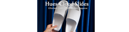 A pair of white Hues Cloud Slides held by two hands in front of a rich blue curtain, showcasing their textured upper strap, cushioned sole, and minimalist slip-on design.